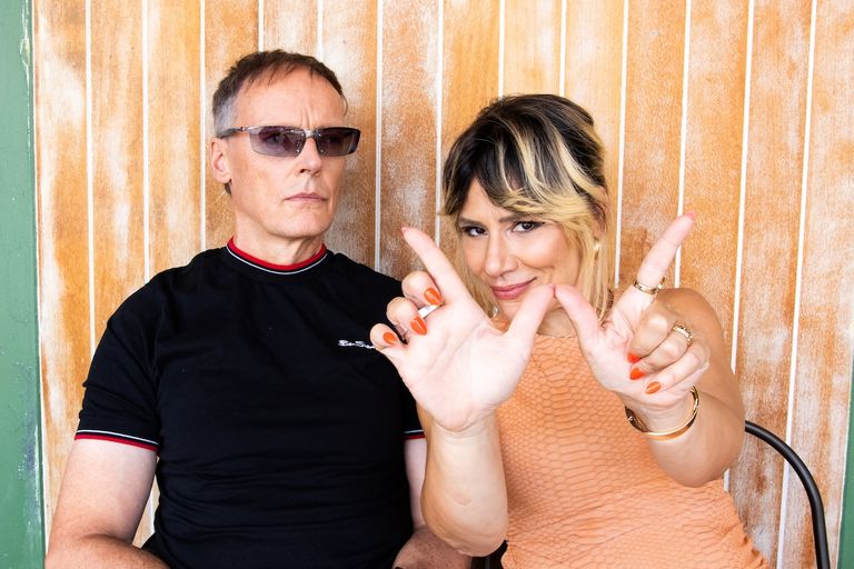 Australian pop duo Bachelor Girl posing together, with Tania Doko making hand signs toward the camera alongside James Roche in sunglasses.